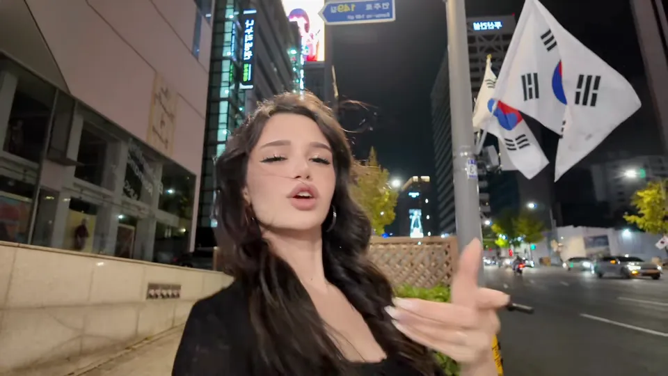 A woman with dark wavy hair points at the camera, Korean flags waving on a pole behind her