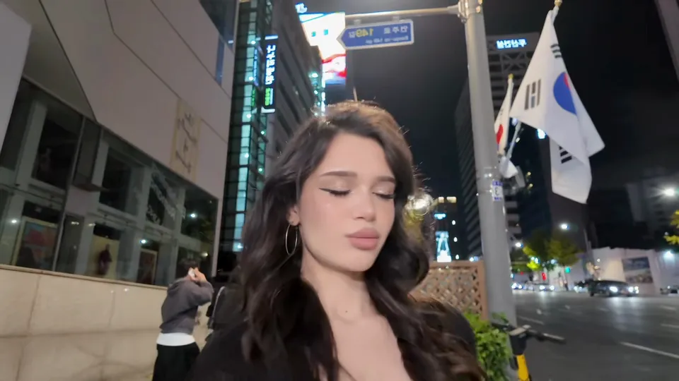 The woman with dark wavy hair sings to the camera under Korean flags on a street at night