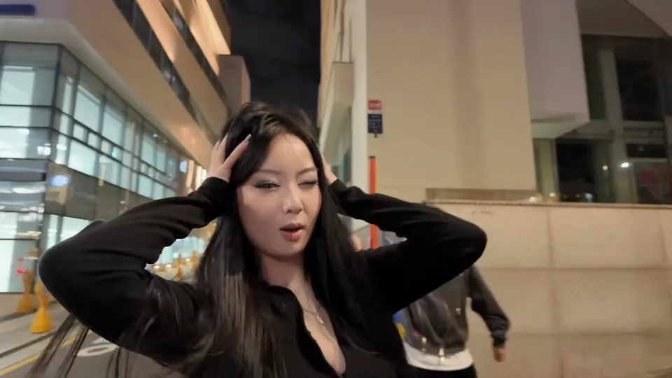 A woman in black runs both hands through her long hair on a pavement beside a glass office block