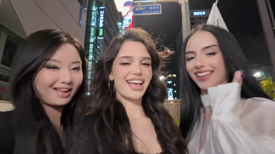 Three friends squeezed into a night selfie on a Seoul street, tall buildings and a lit sign behind them