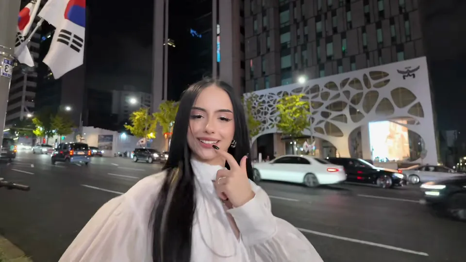 A woman in a white blouse touches her chin by a busy road at night, a lattice-front building behind her